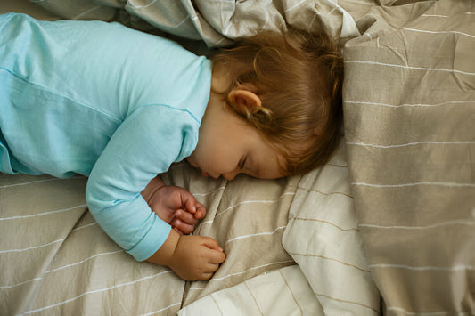 Bebé niña dormida en una cama con sábanas color beige y crema. La bebé está vestida de color azul