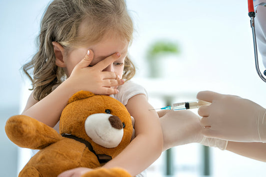 Niña pequeña tapandose los ojos y sosteniendo un oso de peluche mientras un médico está por vacunar a la bebé